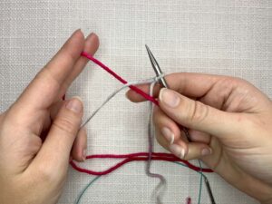 Provisional Cast On Without Crochet Hook: Hold your needle in your right hand, take the tail of the scrap yarn and place it between the working yarn tail and your needle, pinching it with your thumb and middle finger to keep it in place.