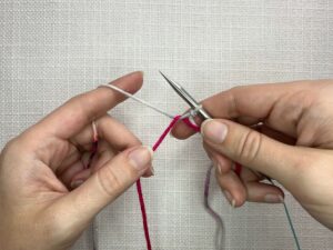 Provisional Cast On Without Crochet Hook: Holding the scrap yarn in your left hand, take the working yarn and place it at the back of the scrap yarn and up and over the front of the needle.