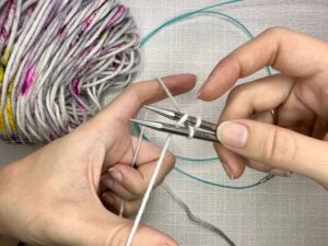 Provisional Cast On Without Crochet Hook: Forming a V with your fingers, hold your yarn tail over your thumb, below both needles, and have your working yarn over your pointer finger, above both needles.